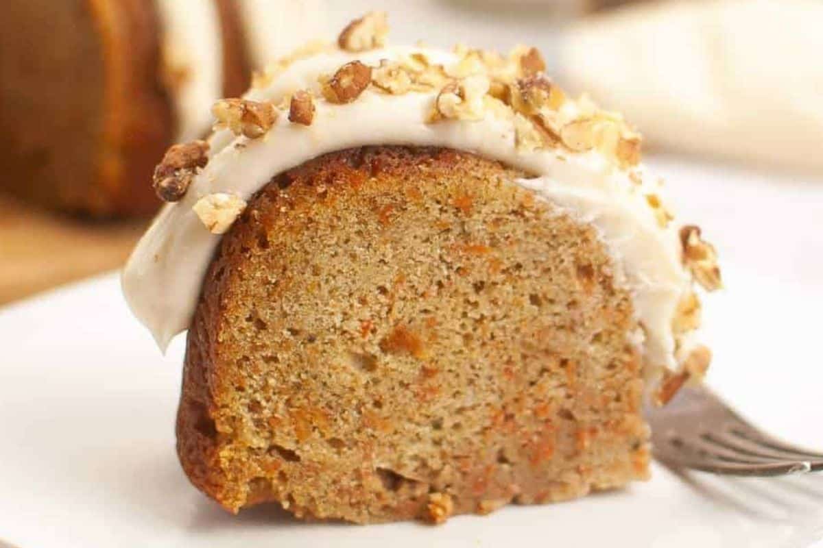 A slice of irresistible carrot Bundt cake, inspired by a classic Nothing Bundt Cake recipe, is adorned with creamy cheese frosting and a sprinkle of chopped nuts. It sits elegantly on a white plate, waiting beside a fork for your indulgence.