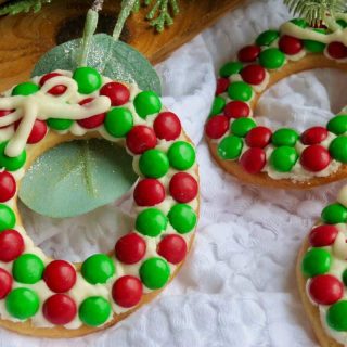 Cookies decorated like wreaths with red and green candies and white icing bows on a white surface.