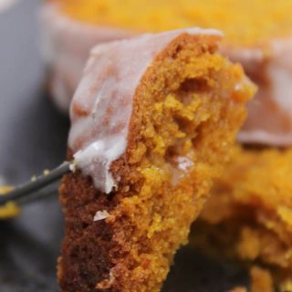 Close-up of a slice of pumpkin loaf with a glaze, showing a partly golden-brown crust and soft interior. A fork is gently lifting a piece.