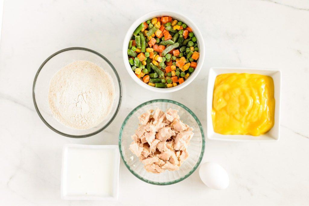 Ingredients on a countertop: flour, mixed vegetables, cream of chicken soup, milk, shredded chicken, and an egg.