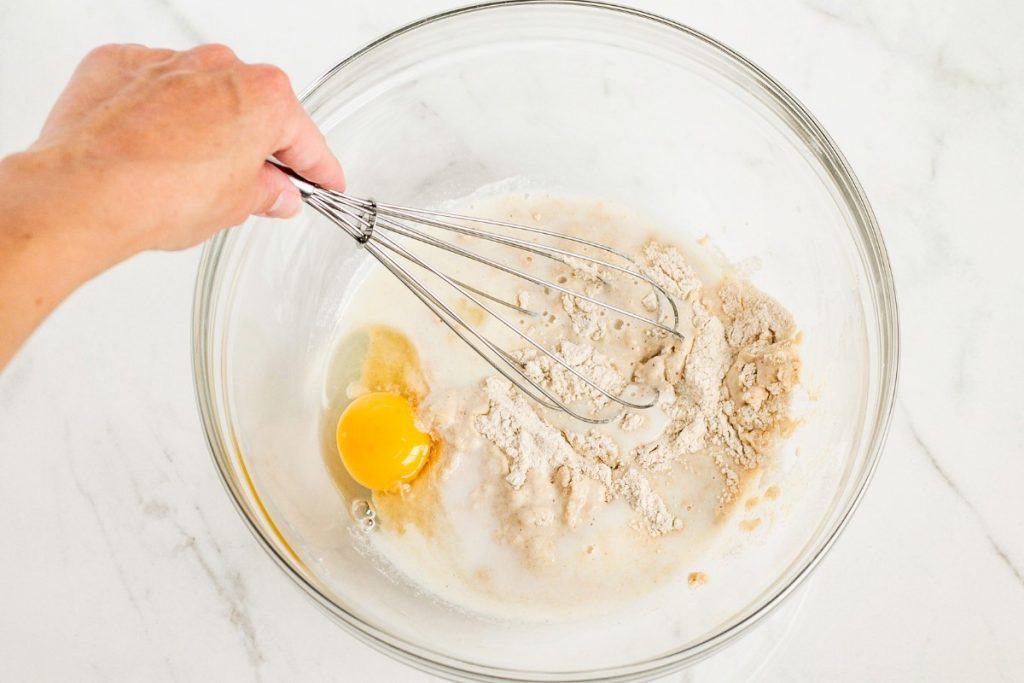 A hand holds a whisk over a glass bowl containing an egg, flour, and liquid ingredients, ready for mixing on a white surface.