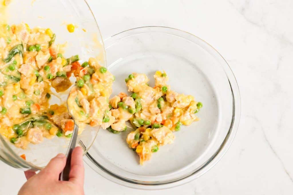 A person is pouring a mixture of chicken, peas, corn, and carrots into a glass pie dish on a marble surface.