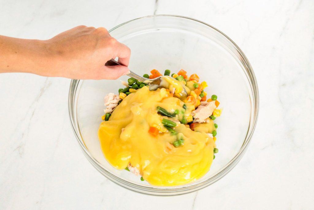 A hand mixes vegetables, meat, and yellow sauce in a glass bowl with a spoon.