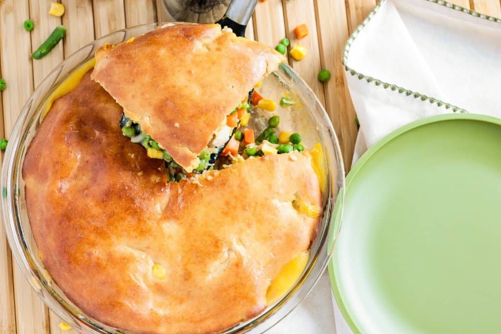 A pot pie with a golden crust is partially cut open, revealing vegetables inside. It sits on a wooden surface next to a green-rimmed plate.
