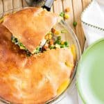 A pot pie with a golden crust is partially cut open, revealing vegetables inside. It sits on a wooden surface next to a green-rimmed plate.