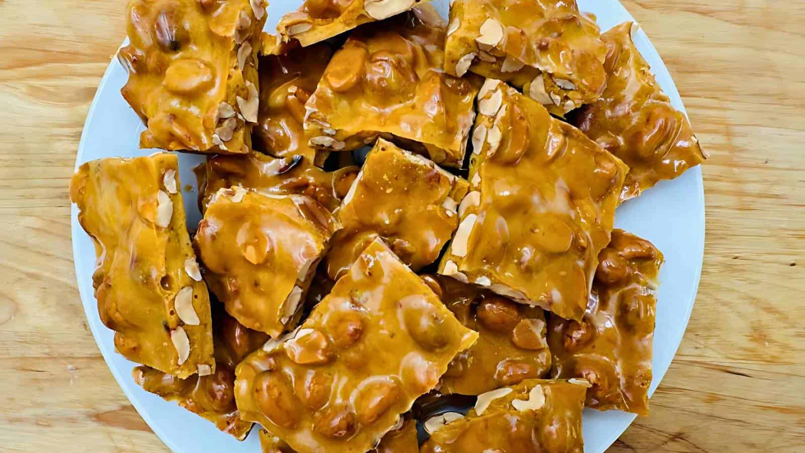A plate of golden brown peanut brittle pieces on a wooden surface.