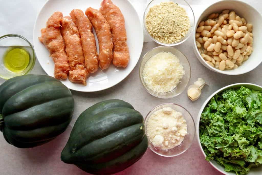 Ingredients on a table: raw sausages, breadcrumbs, white beans, olive oil, acorn squash, grated cheese, garlic clove, and fresh kale.