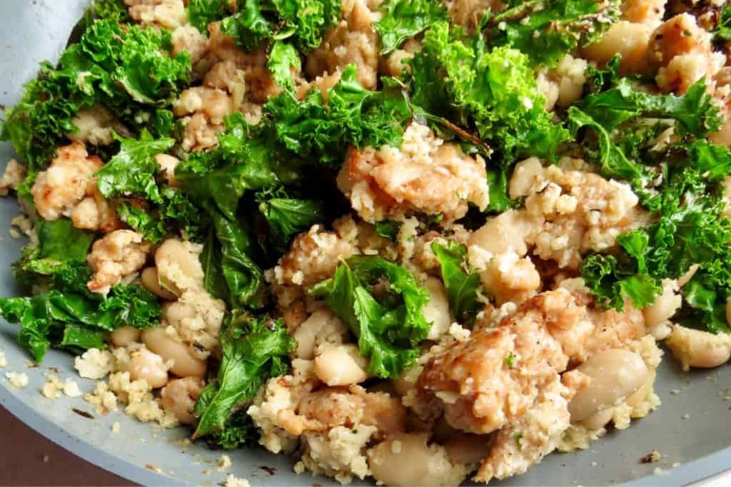 A skillet holds a dish with sautéed kale, white beans, and crumbled sausage.