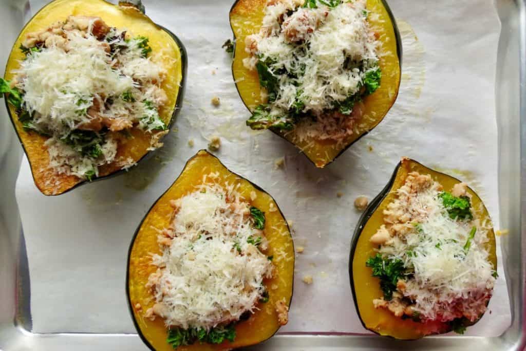 Four halved roasted acorn squash filled with a stuffing of ground meat, greens, and topped with grated cheese on a baking sheet.