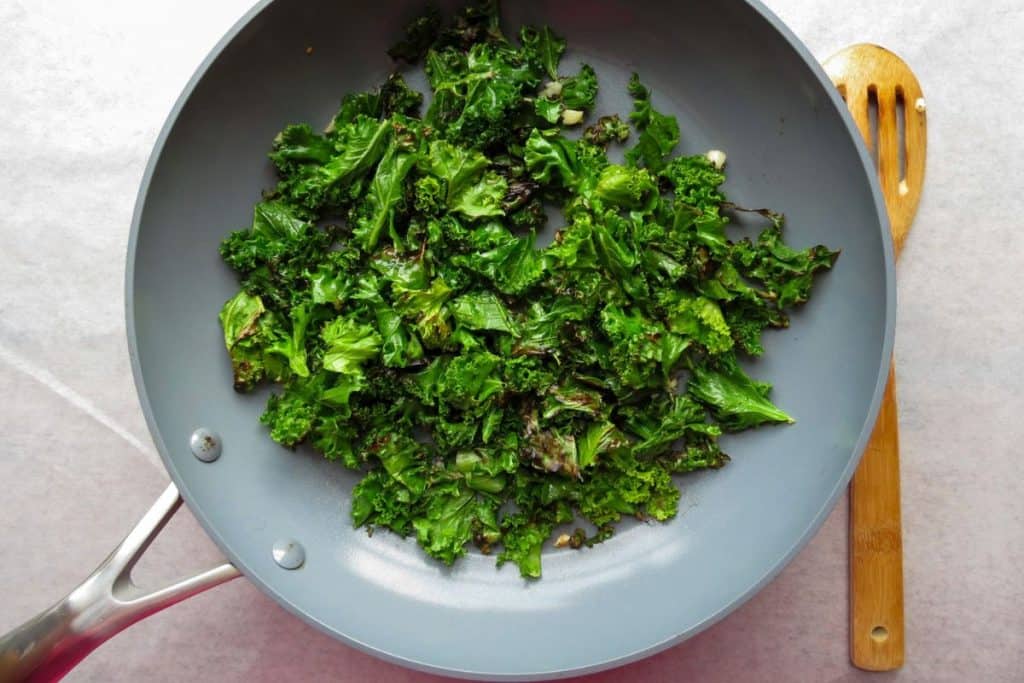 A frying pan with cooked kale on a light surface, next to a wooden spoon.