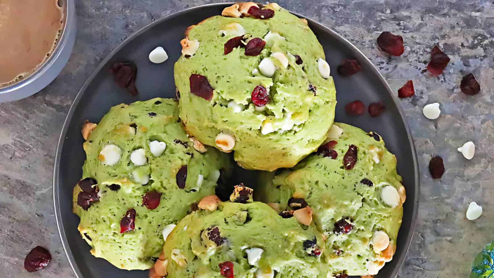 Green cookies with white chocolate chips and dried cranberries on a dark plate, next to a cup of coffee.