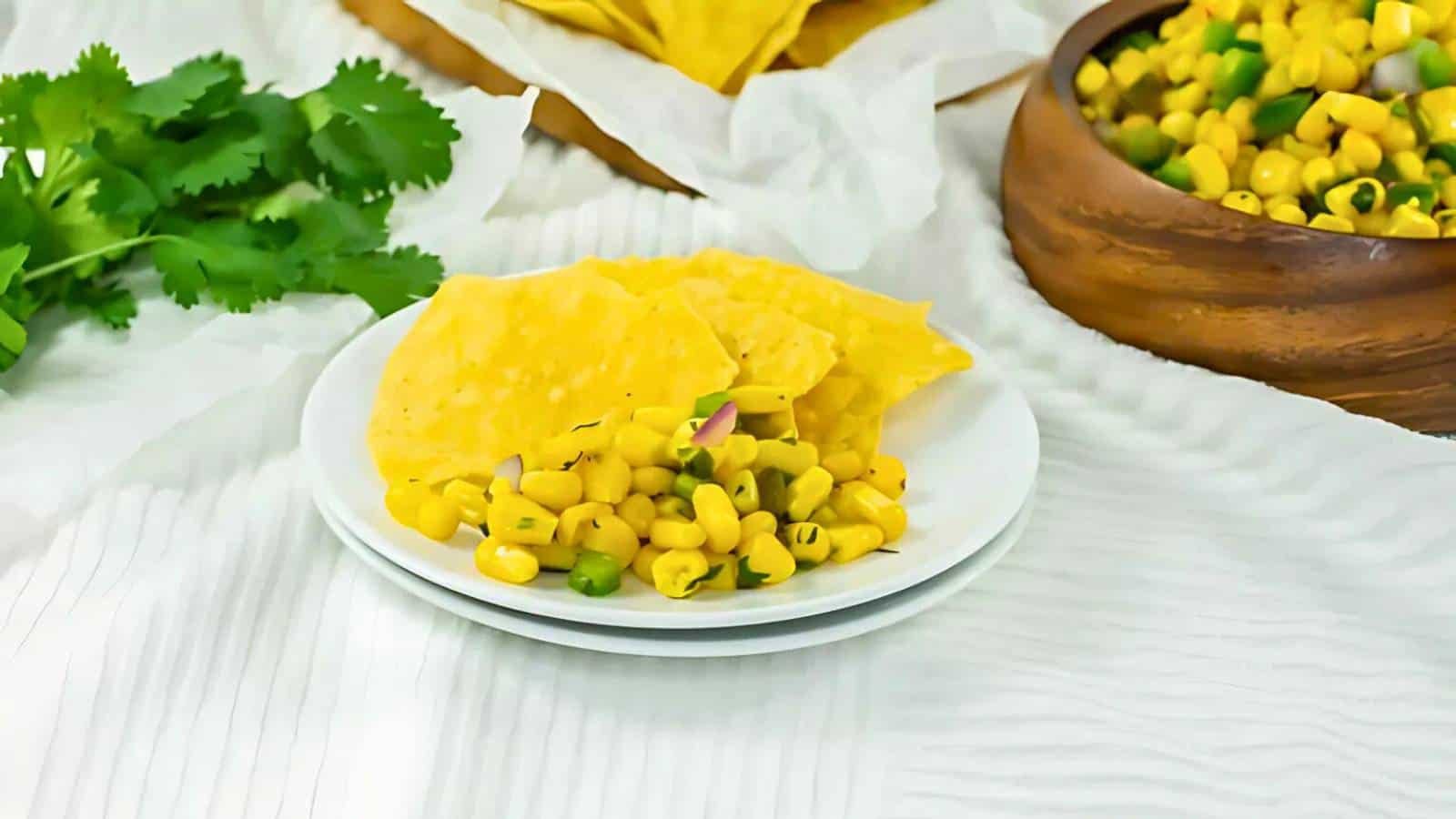 White plates with corn salad and tortilla chips, surrounded by a wooden bowl of corn salad and fresh cilantro on a white cloth.
