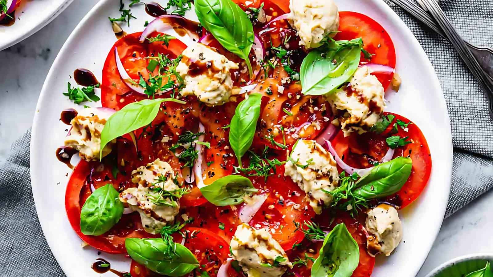 A plate of sliced tomatoes and red onions topped with dollops of cheese, fresh basil leaves, herbs, and drizzled with balsamic glaze.