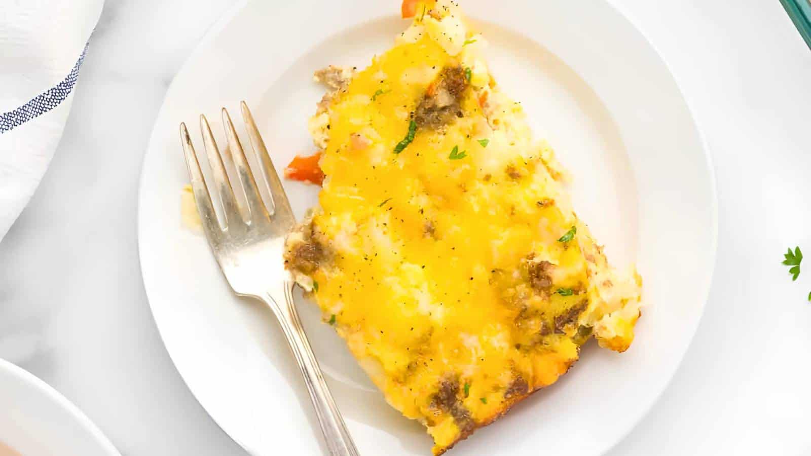 Slice of casserole with melted cheese and ground meat on a white plate, accompanied by a fork.