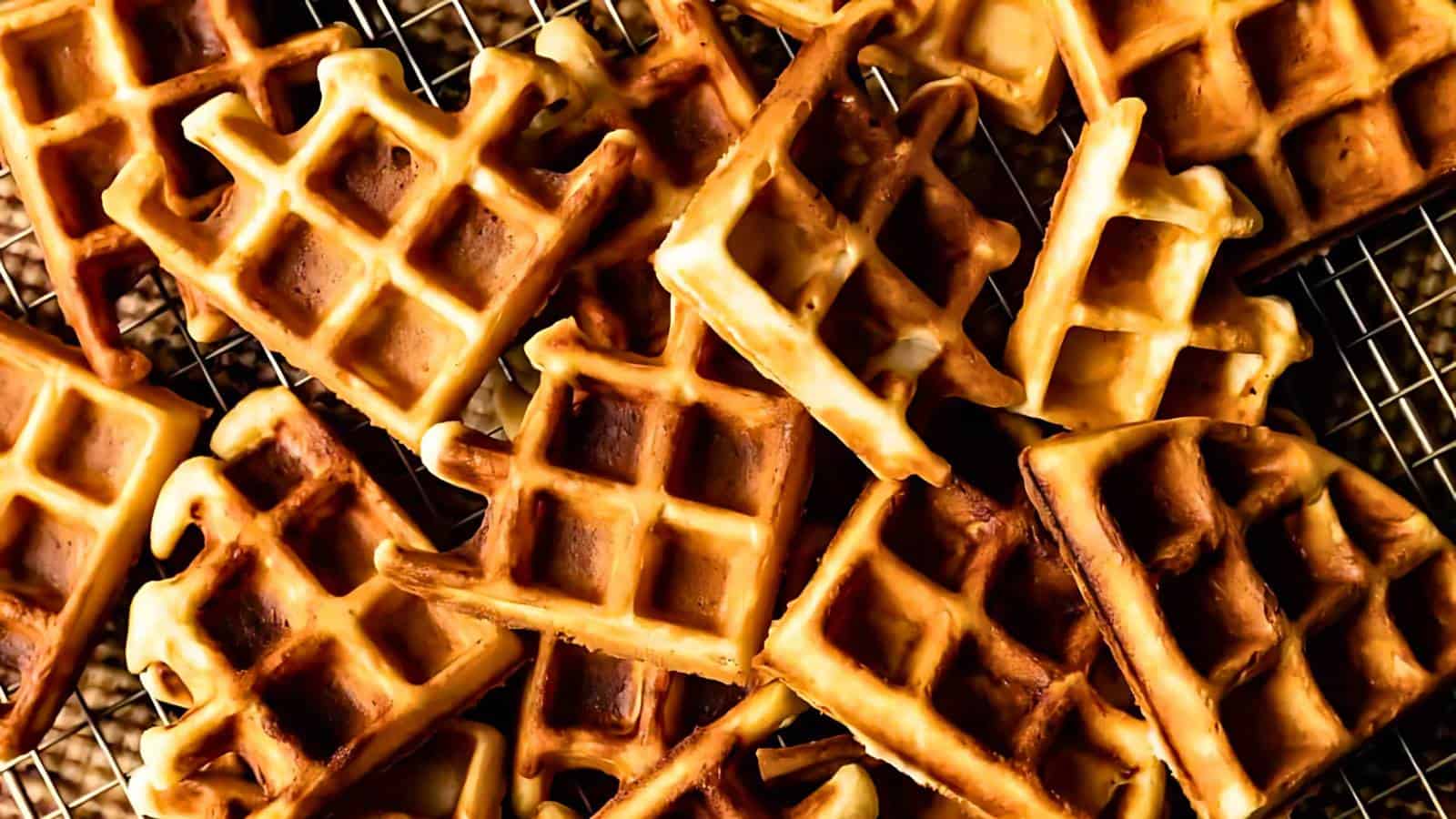 A pile of golden-brown waffles on a cooling rack, showcasing a grid pattern and varied shades of crispiness.