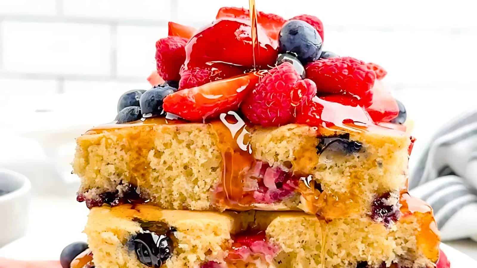 Close-up of stacked pancakes topped with sliced strawberries, raspberries, and blueberries, drizzled with syrup.