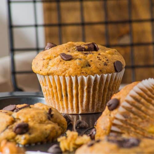 A close-up of peanut butter banana chocolate chip muffins, some wrapped in paper liners, on a baking tray. A cooling rack and wooden board are in the background.