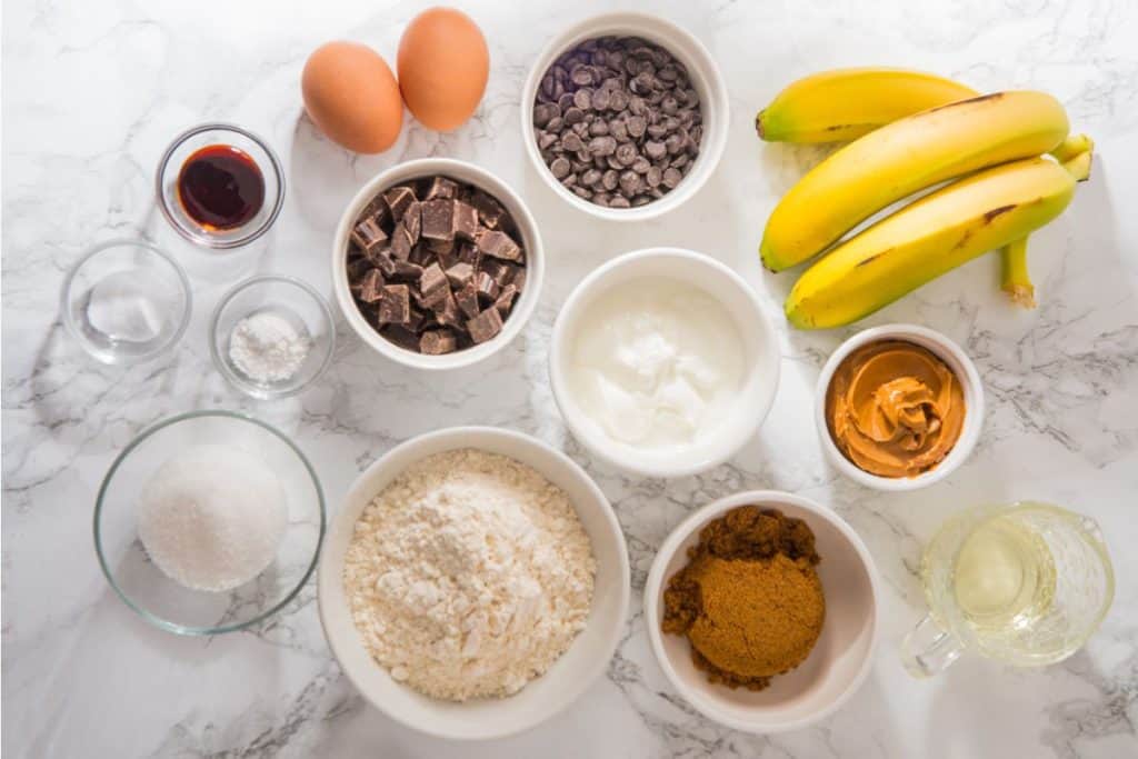 Ingredients for baking include eggs, chocolate chips, bananas, vanilla, baking soda, salt, sugar, flour, yogurt, peanut butter, brown sugar, and oil on a marble surface.