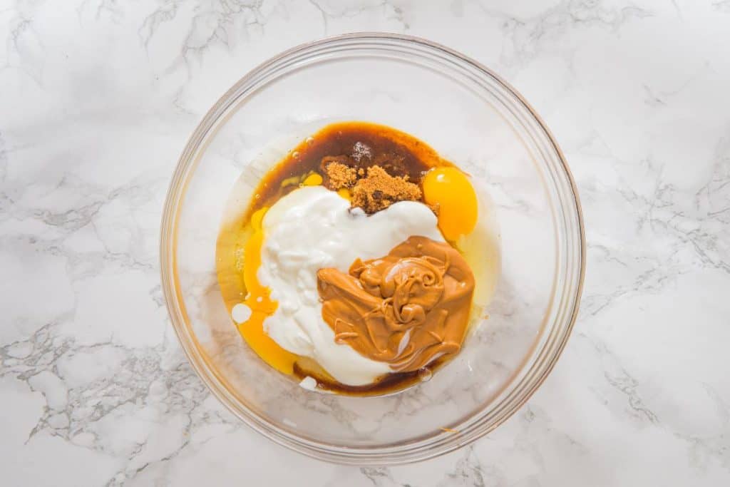 Ingredients in a glass bowl: eggs, peanut butter, yogurt, brown sugar, and vanilla extract on a marble surface.