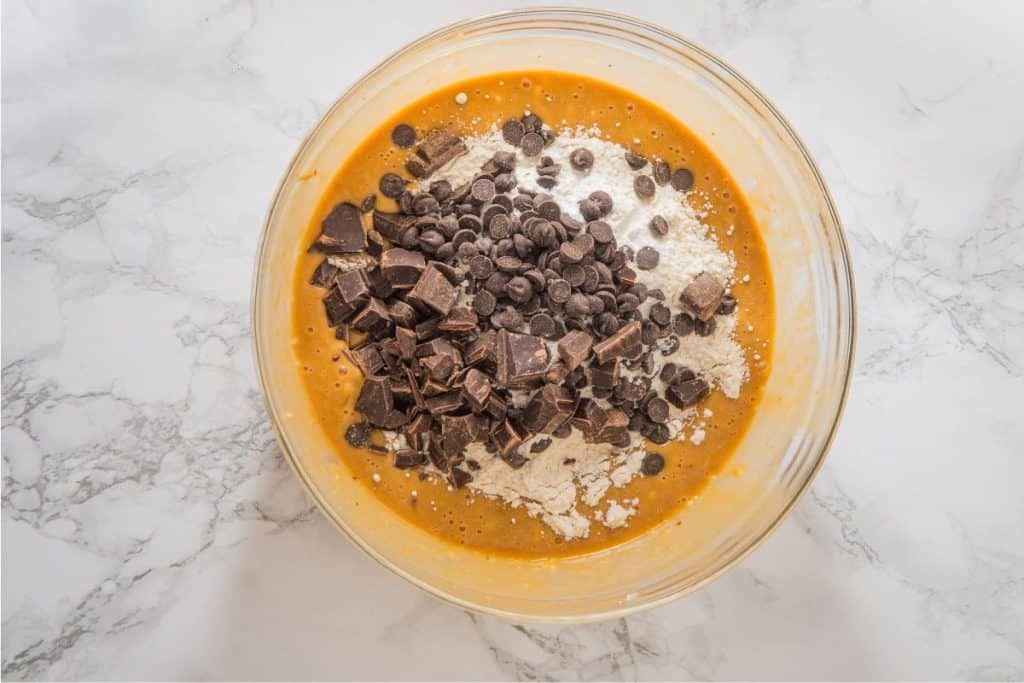 Bowl with cookie dough mixture, containing chocolate chips, chunks, and flour, on a marble countertop.