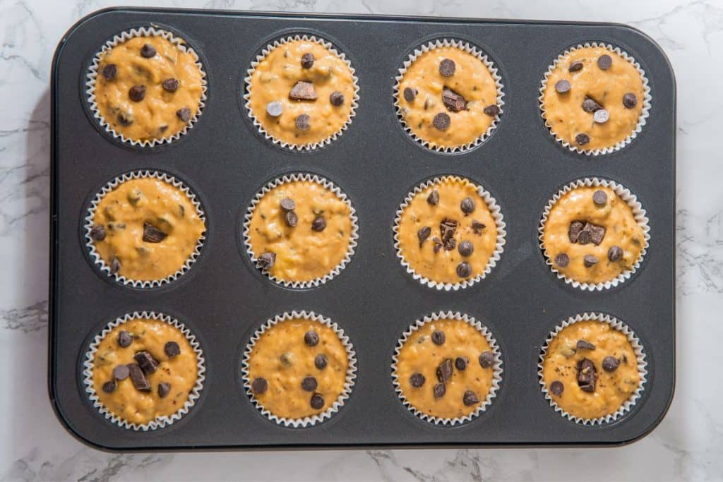 Muffin tray with twelve unbaked muffins, each with chocolate chips and chunks, on a marble countertop.