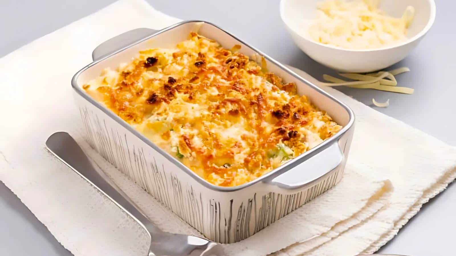 Baked pasta dish with melted cheese in a rectangular dish on a towel, next to a metal spatula and a bowl of shredded cheese.