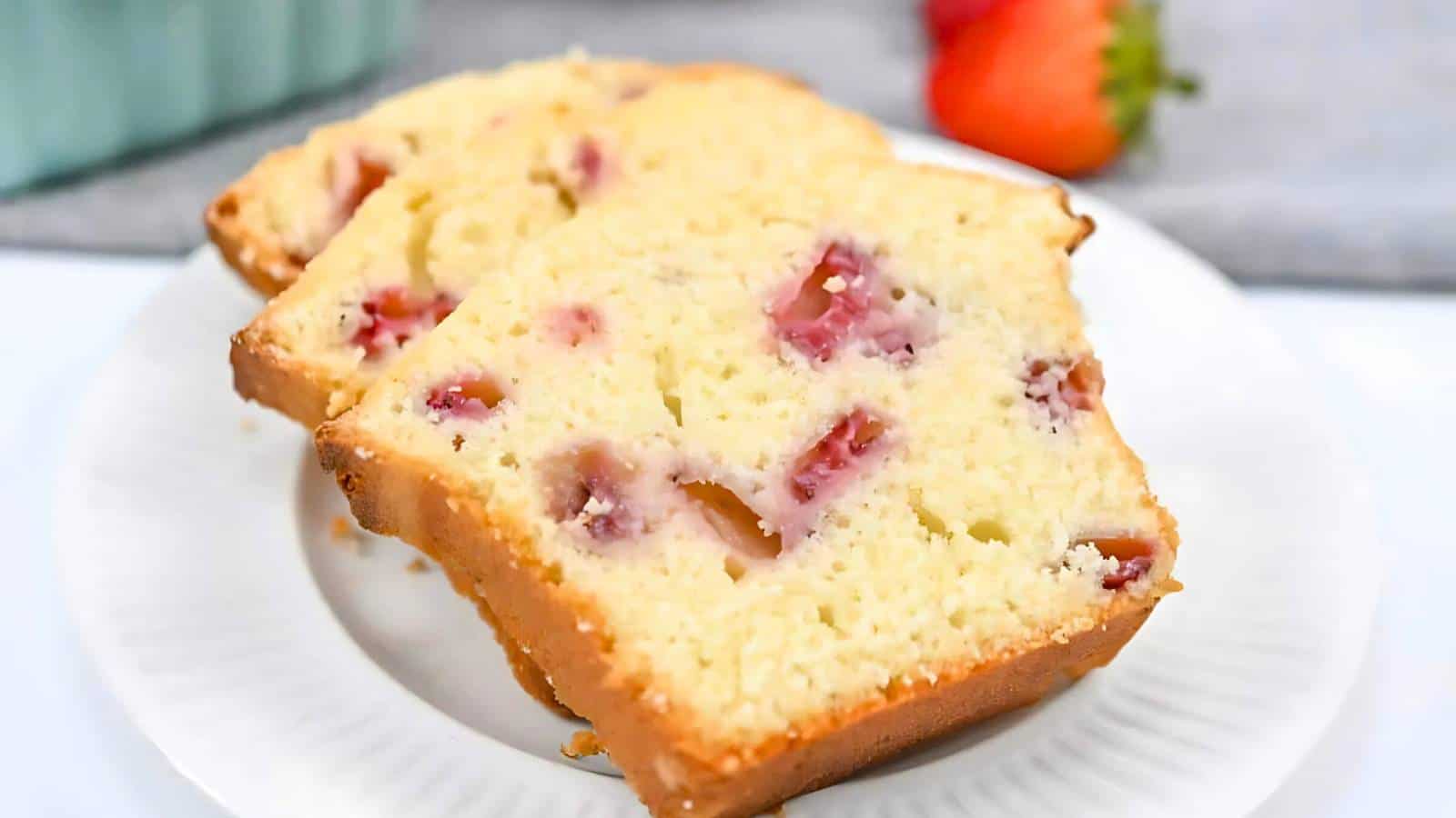 Two slices of strawberry bread are on a white plate, with visible strawberry pieces. A strawberry is blurred in the background.