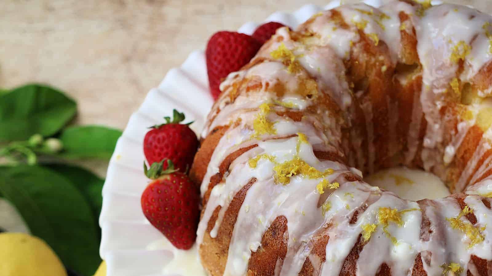 Bundt cake with white icing and lemon zest on a white plate, garnished with fresh strawberries.