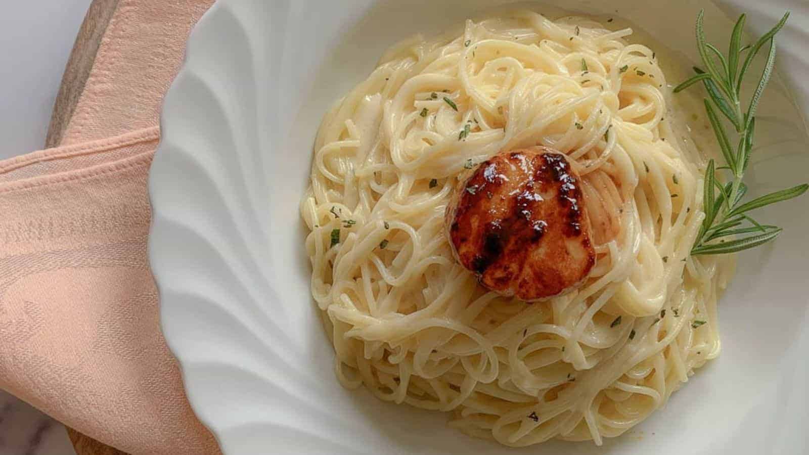 A plate of creamy pasta topped with a seared scallop, garnished with a sprig of rosemary.