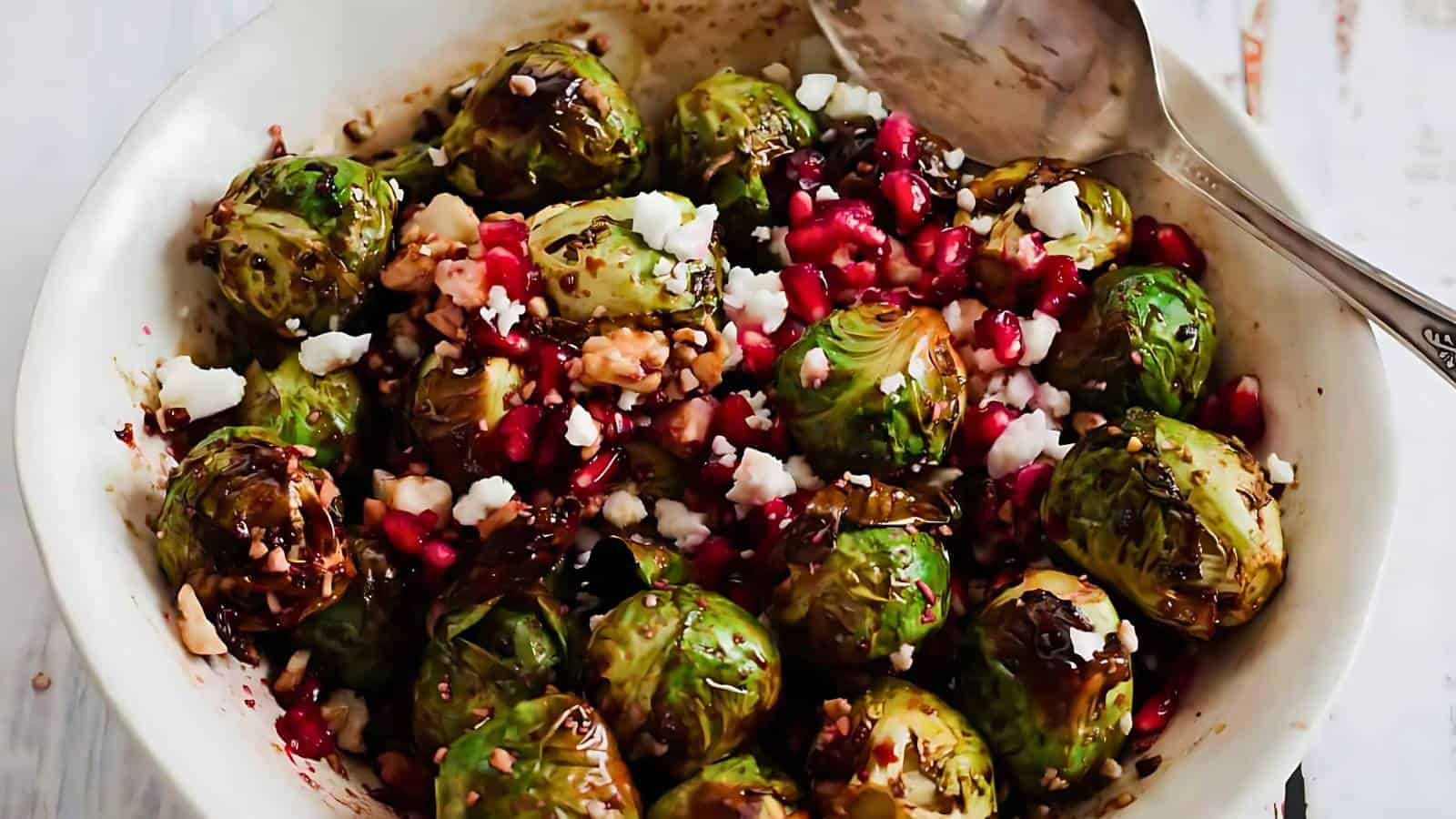 A bowl of roasted Brussels sprouts with pomegranate seeds, walnuts, and crumbled feta cheese, served with a spoon.