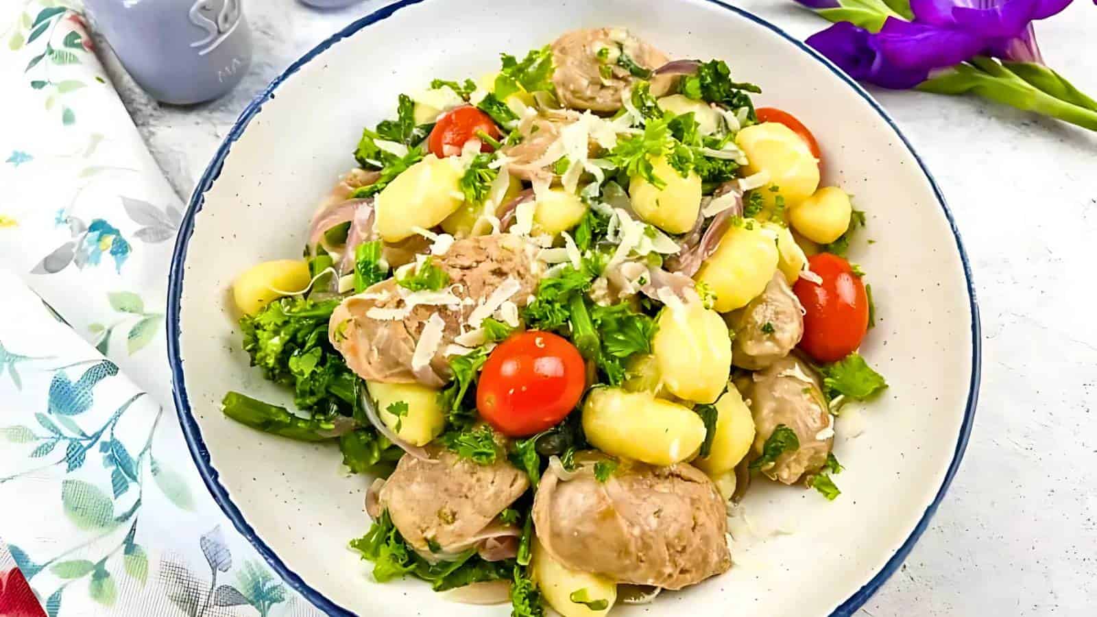 A bowl of gnocchi with sausage, cherry tomatoes, broccoli, and grated cheese, garnished with parsley. A floral napkin and purple flower are nearby.