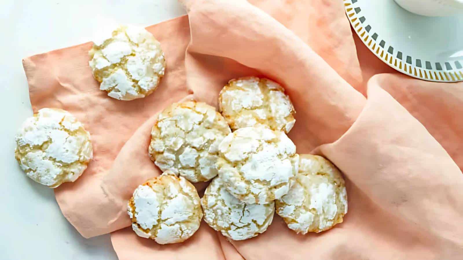 A group of powdered sugar-covered cookies arranged on a pink cloth next to a white plate with a gold rim.