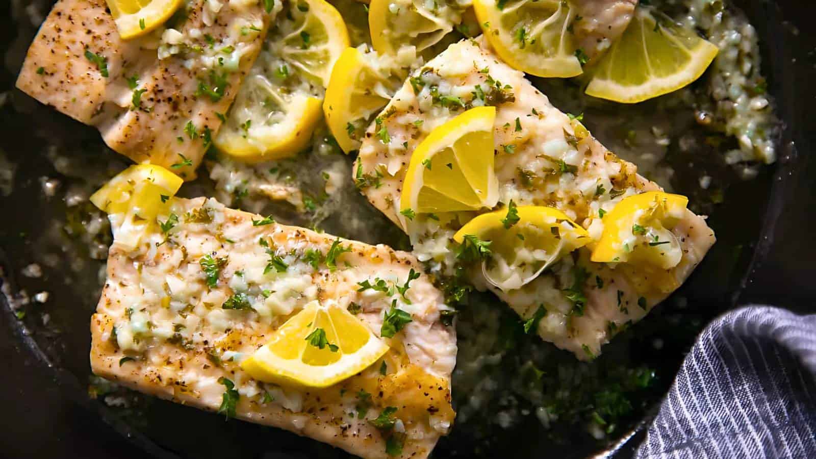 Cooked fish fillets garnished with lemon slices and herbs in a pan, accompanied by a striped cloth.