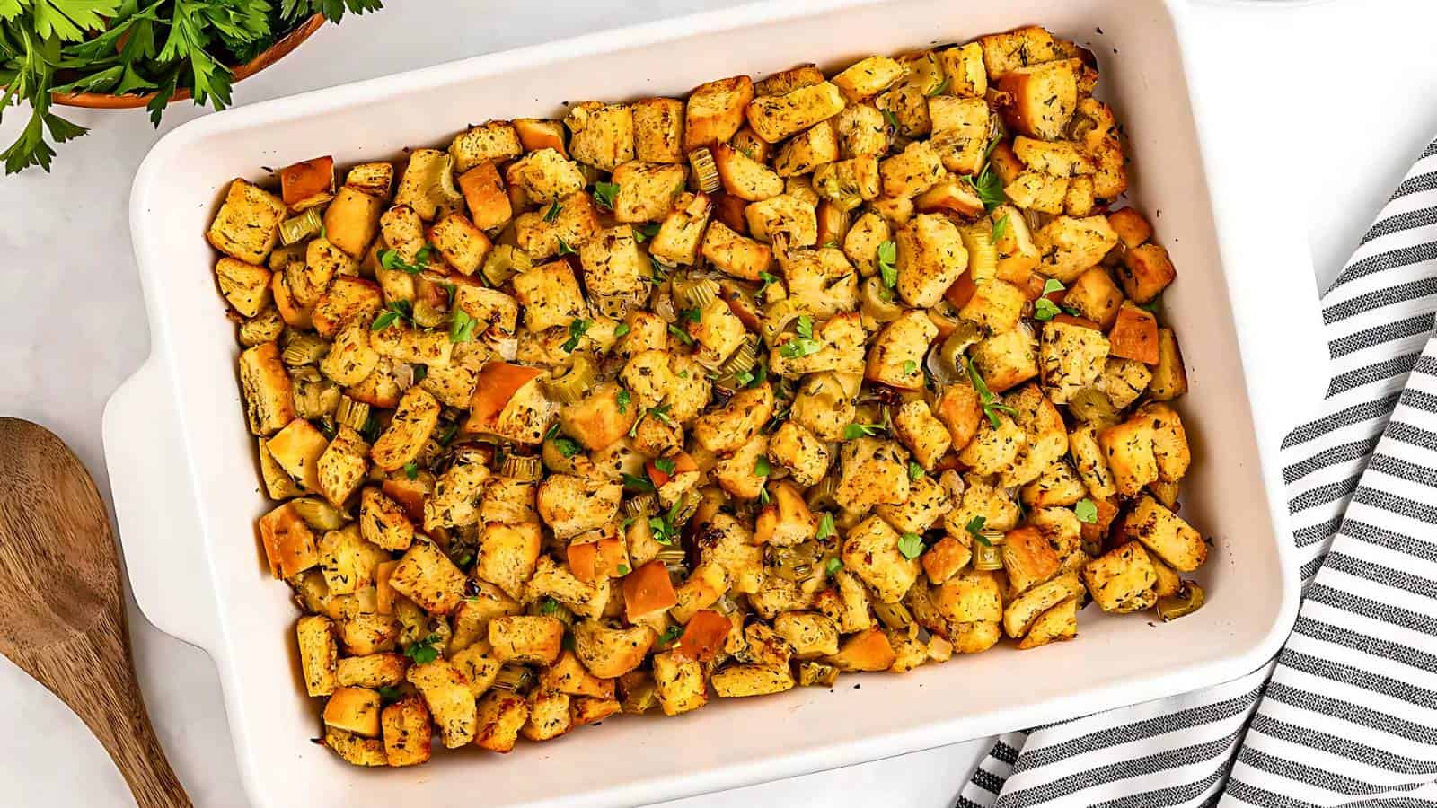 A white casserole dish filled with seasoned croutons, herbs, and diced vegetables, on a striped cloth with a wooden spoon nearby.