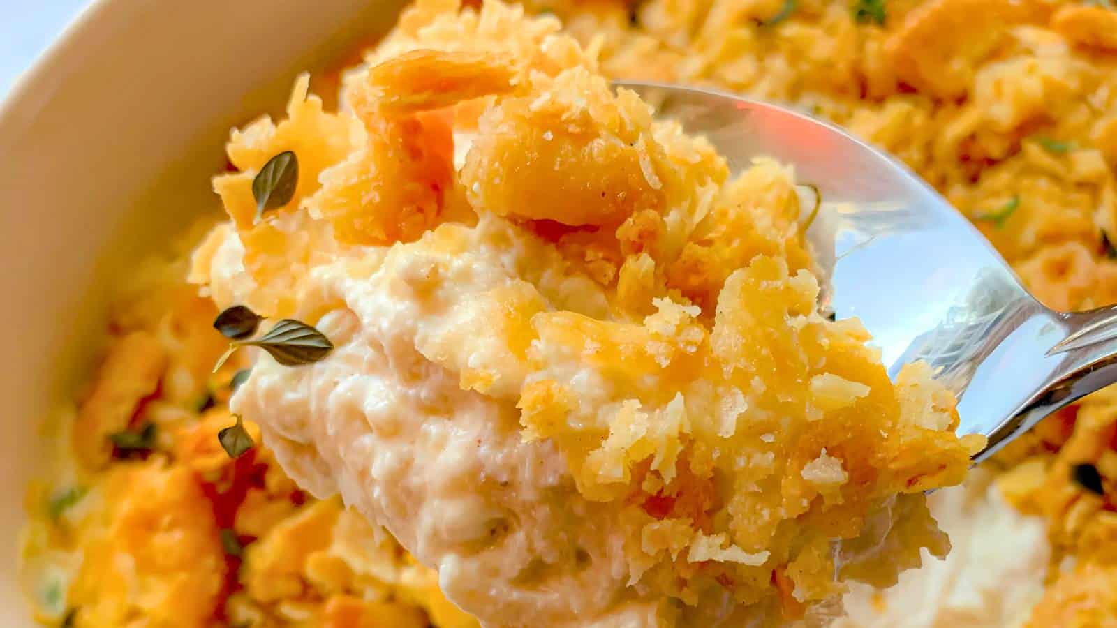 Close-up of a spoonful of cheesy casserole with a crispy topping, garnished with small green herbs, above a dish filled with the casserole.