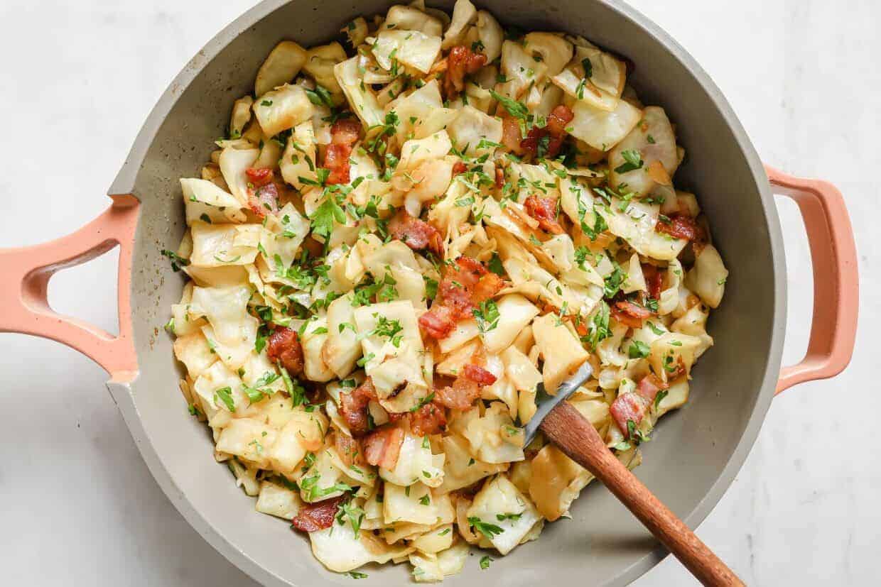 A pan of cooked cabbage and bacon garnished with parsley, with a wooden spoon.