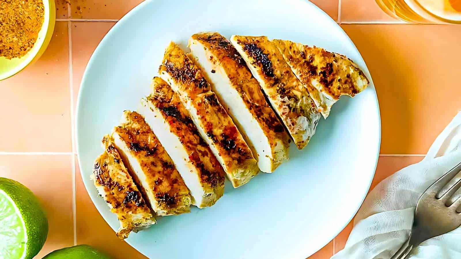 A sliced grilled chicken breast on a white plate, garnished with lime wedges and accompanied by a napkin and utensils.