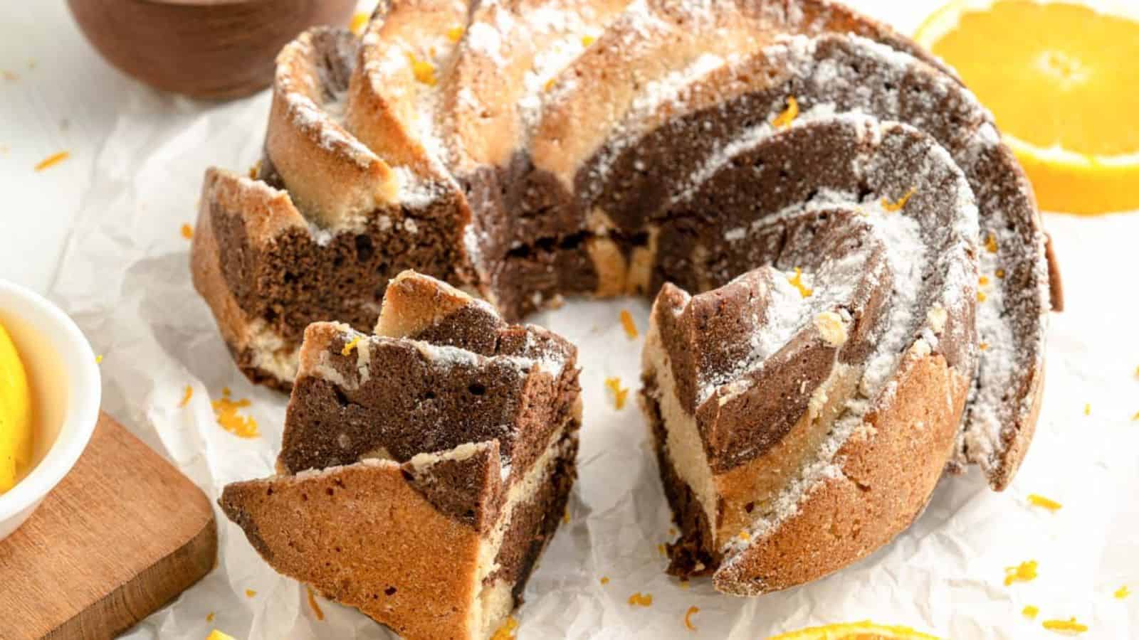 Marble bundt cake with powdered sugar on parchment paper, surrounded by orange slices.