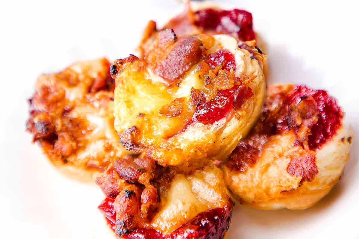 Four baked pastry bites with melted cheese, crispy bacon bits, and red jam on a white plate.