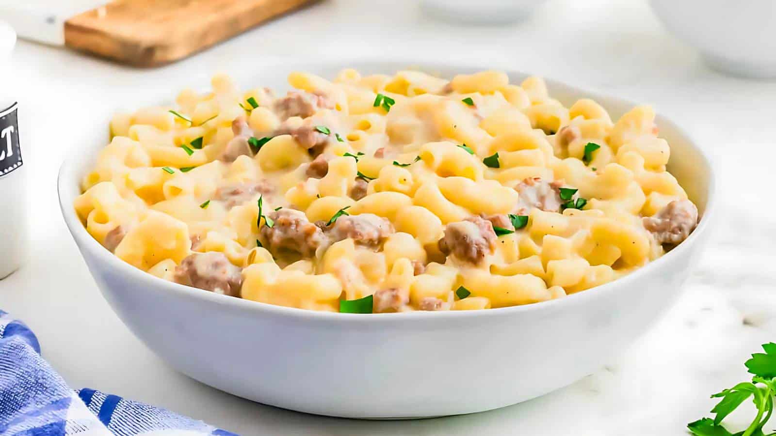 A white bowl filled with creamy macaroni and cheese with ground meat, garnished with chopped herbs, on a white surface.