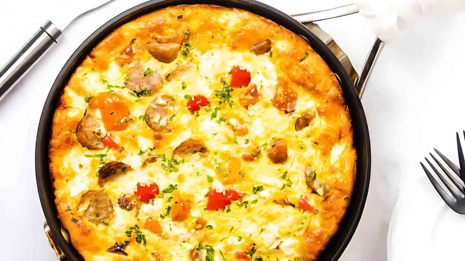 A cooked frittata with visible chunks of sausage, pieces of tomatoes, and garnished with herbs, placed in a black pan on a white background near utensils.