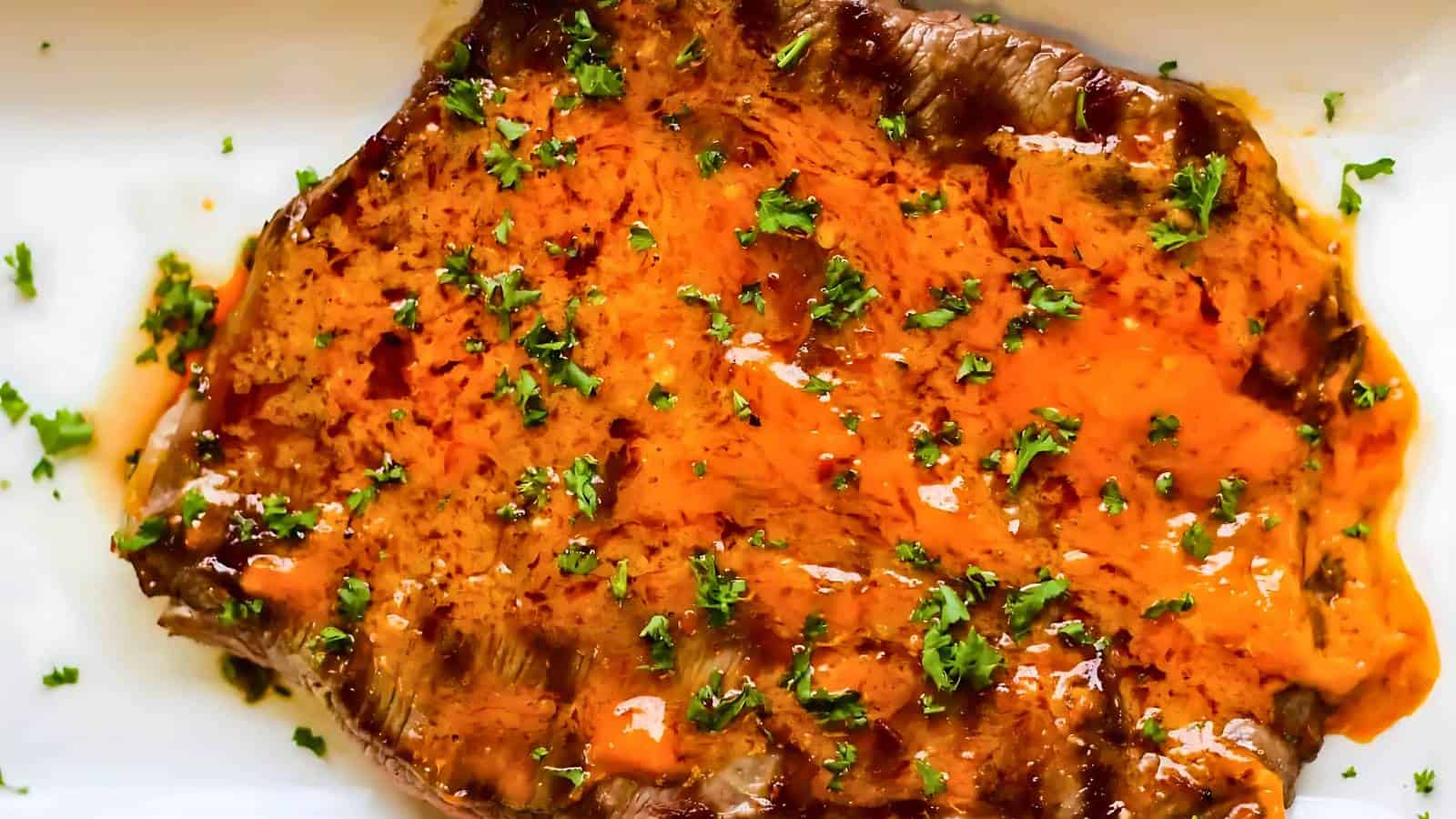 A grilled steak topped with orange sauce and garnished with chopped parsley on a white plate.