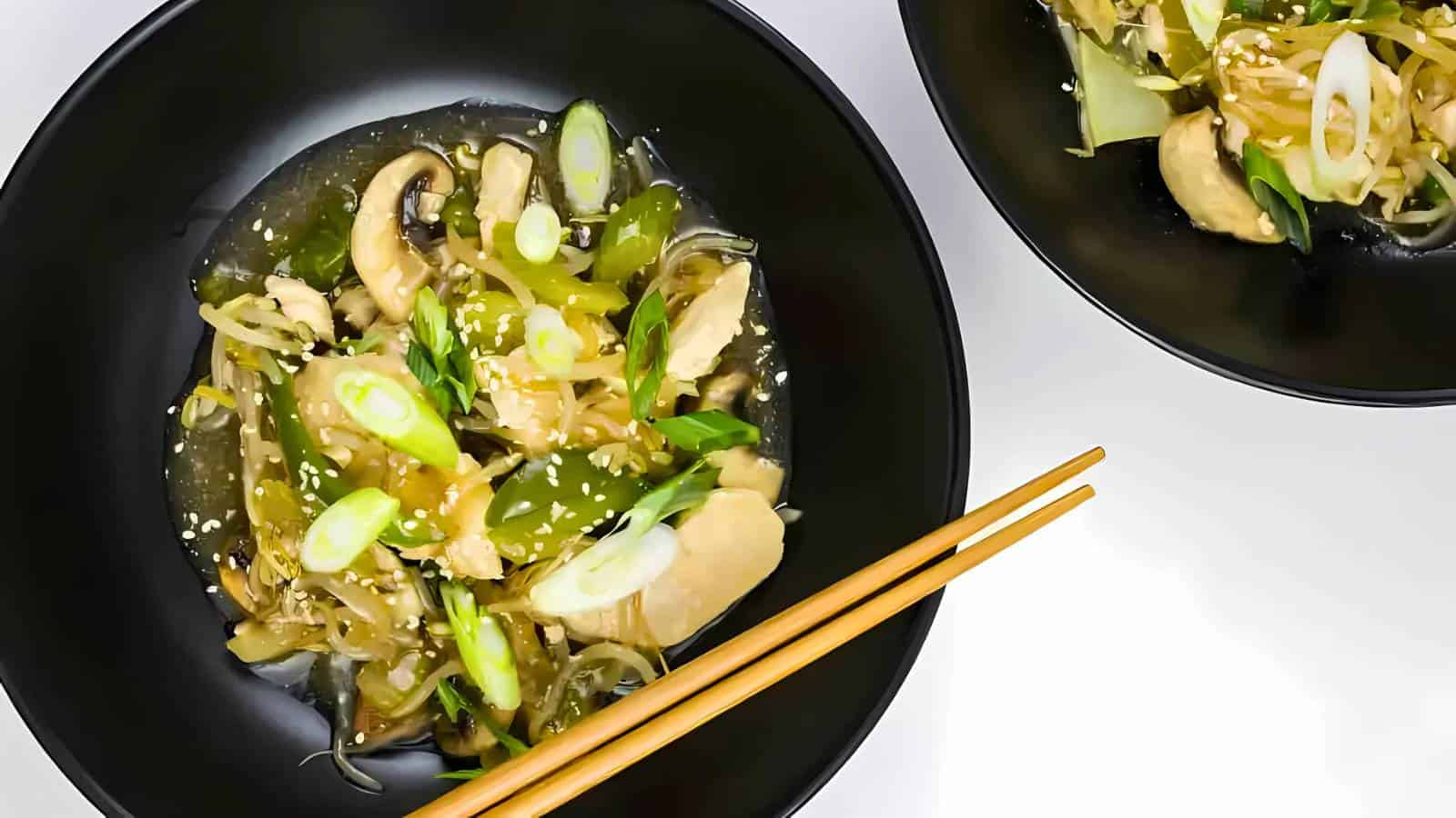 Two black bowls of stir-fried chicken with vegetables, topped with sesame seeds and green onions. Chopsticks are placed on the rim of one bowl.