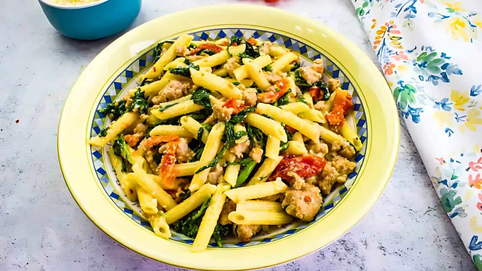 A plate of penne pasta mixed with sausage, wilted greens, and cherry tomatoes, served on a yellow-rimmed dish. A floral napkin is partially visible on the right side.