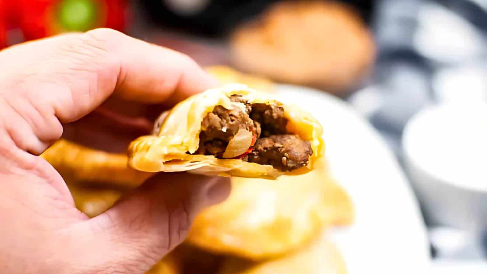Hand holding a half-eaten empanada filled with seasoned meat and vegetables, with a blurred plate of more empanadas in the background.