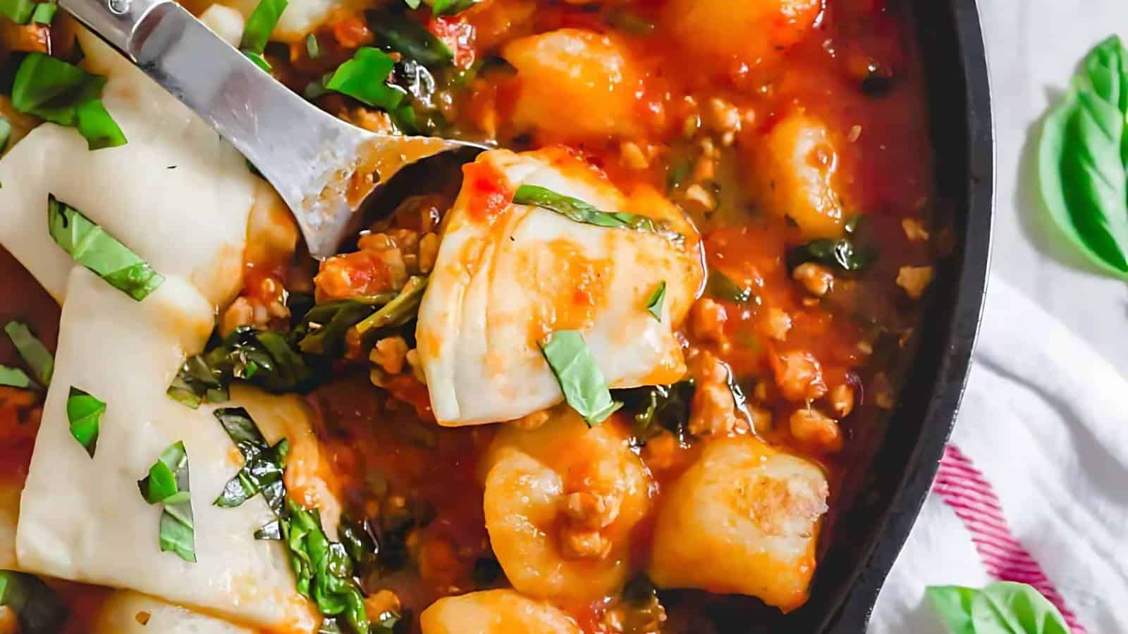 Close-up of a skillet with cooked cheese-stuffed pasta shells, gnocchi, and chopped basil in a tomato sauce. A serving spoon is lifting a portion.
