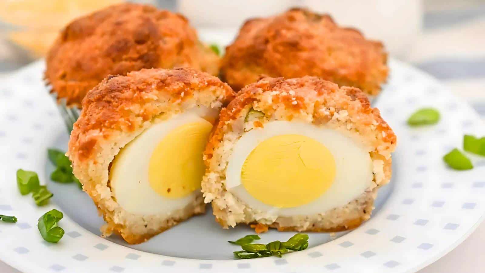 Two scotch eggs on a plate with one cut in half, revealing a boiled egg inside. Scattered green garnish surrounds the eggs.
