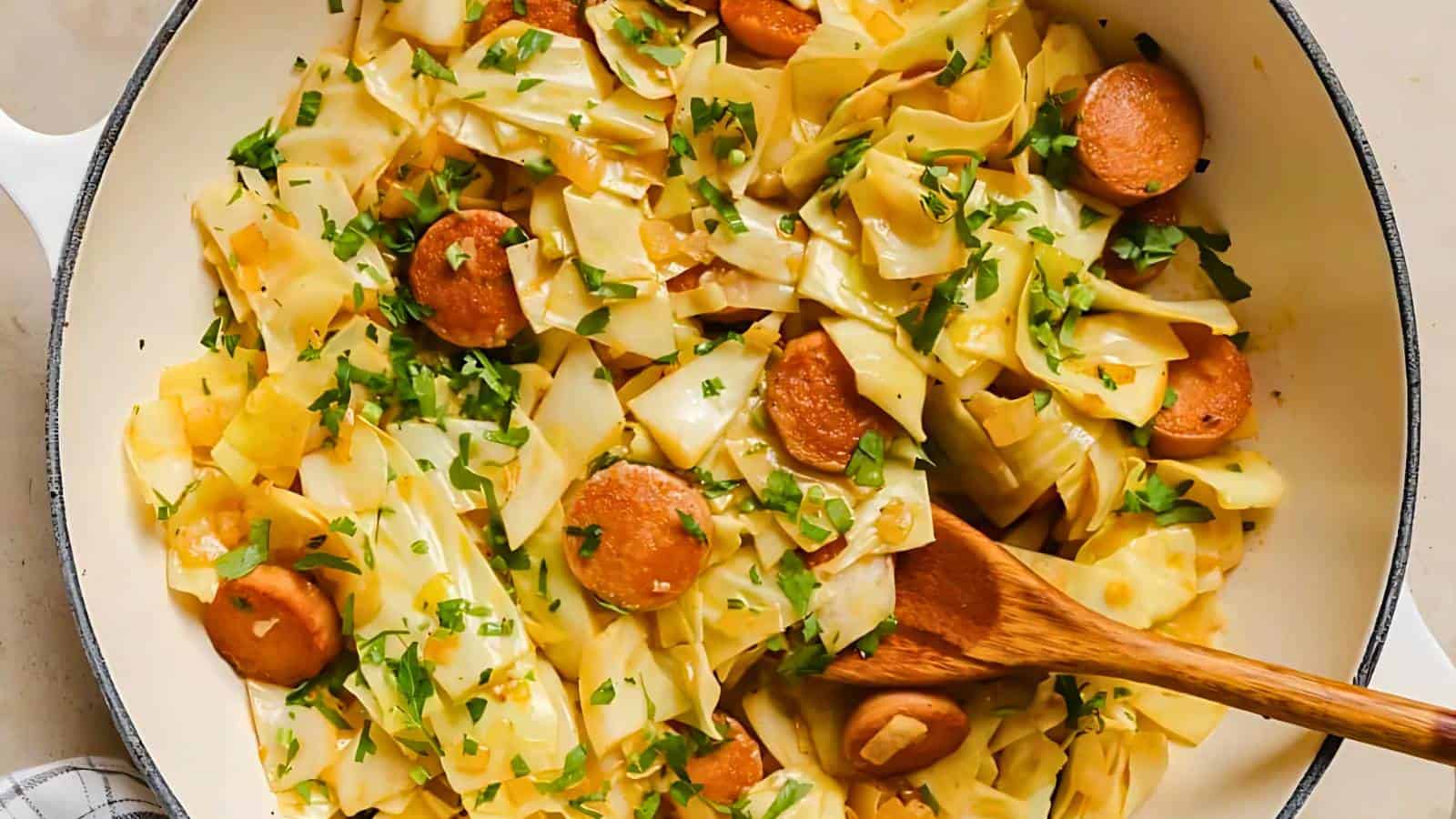 A skillet contains cooked cabbage, sliced sausages, onions, and chopped parsley, mixed together with a wooden spoon.