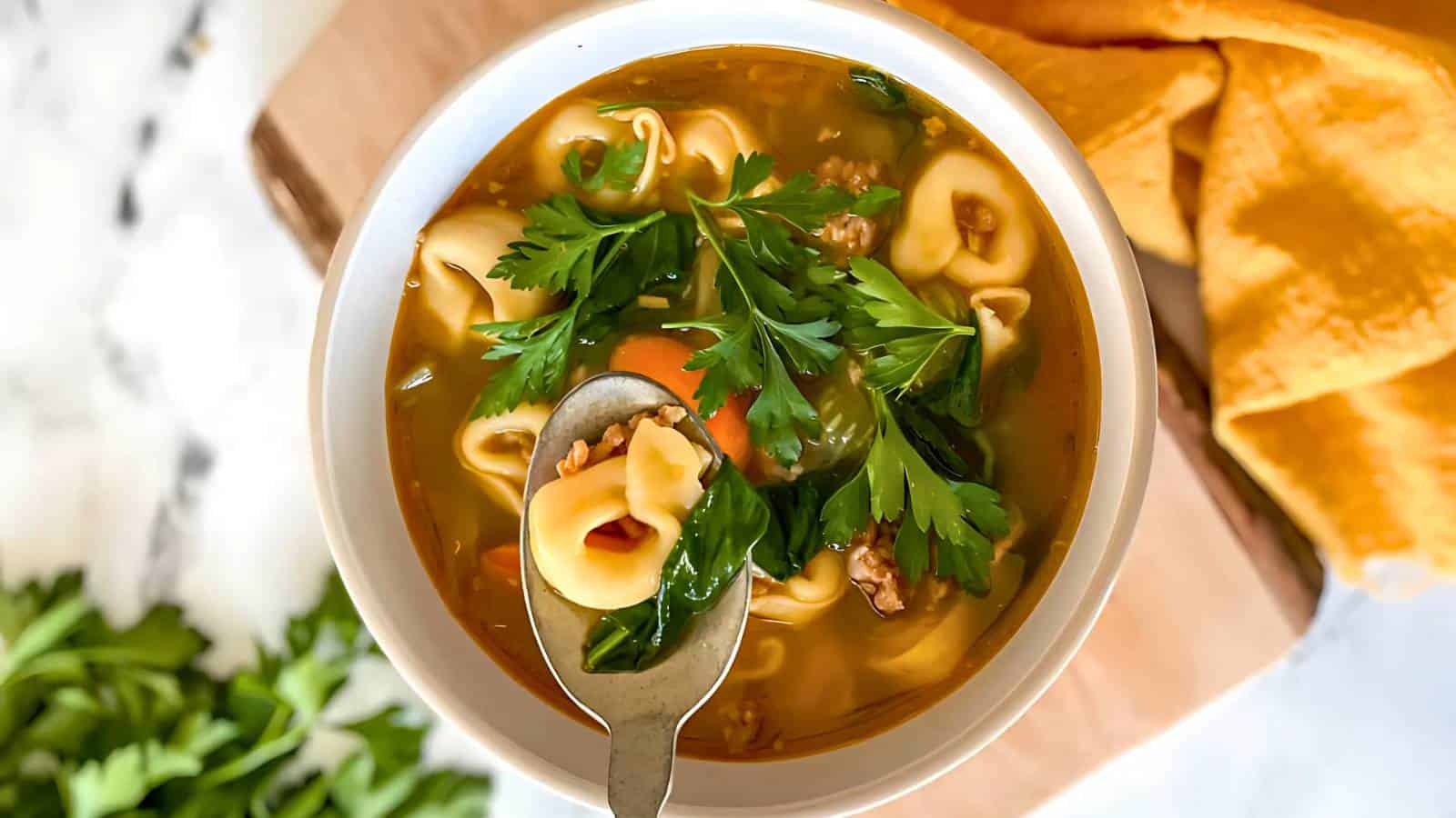 A bowl of tortellini soup with parsley garnish and a spoon, placed on a wooden board beside tortilla chips.