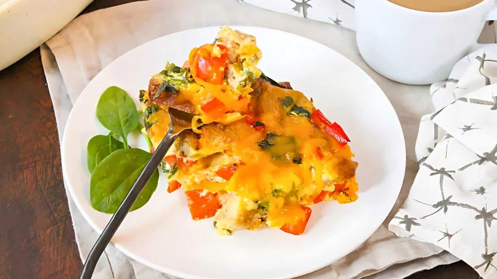 A plate of cheesy vegetable casserole with red peppers and spinach, alongside fresh spinach leaves and a fork, set on a wooden table with a cup of coffee and a patterned napkin.